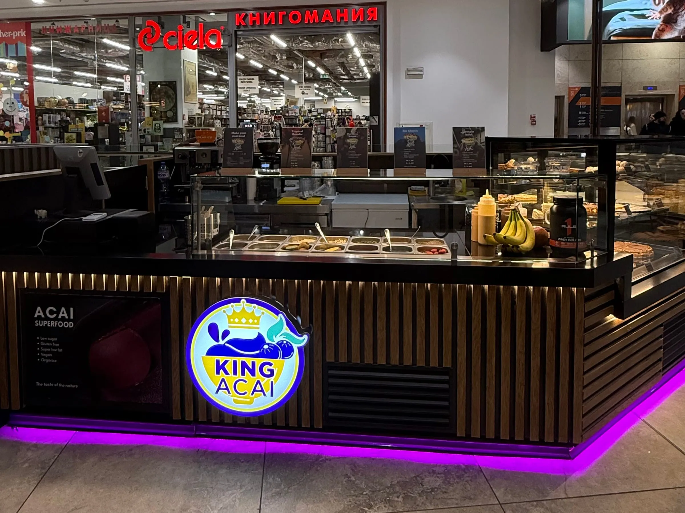 Щандът на King Açaí на ниво 2 в Paradise Center: тъмно дърво, лилава светлина отдолу и логото с короната отпред.